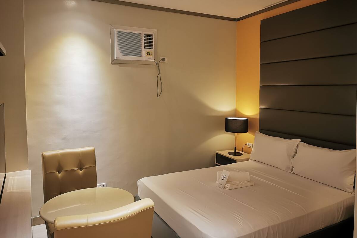 A hotel room with a bed, a chair, a bedside table with a lamp, and an air conditioning unit.