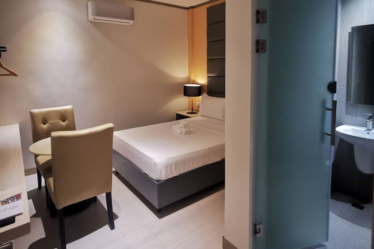 A hotel room with a bed, a chair, a table, an air conditioning unit, a lamp, and a bathroom with a sink and mirror.