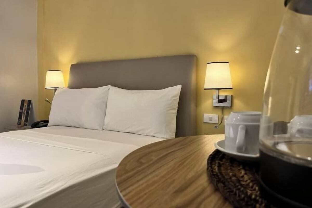 A hotel room with a bed, bedside lamps, a bedside table with a coffee pot, and a wall-mounted phone.