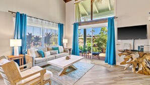 Amplia sala de estar con un sofá grande, una mesa de centro y un televisor. Se observan cortinas azules, una silla de madera y un ventanal con vistas a palmeras.