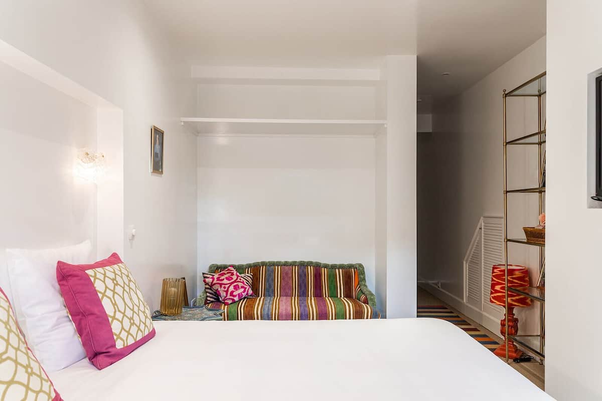 A bedroom with a bed, a colorful striped sofa, a small table, and a shelf with decorative items.
