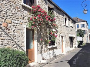 A stone building with a wooden door and windows, a small garden with pink flowers, and a street lamp. - The Rose House (Milly-la-Forêt)