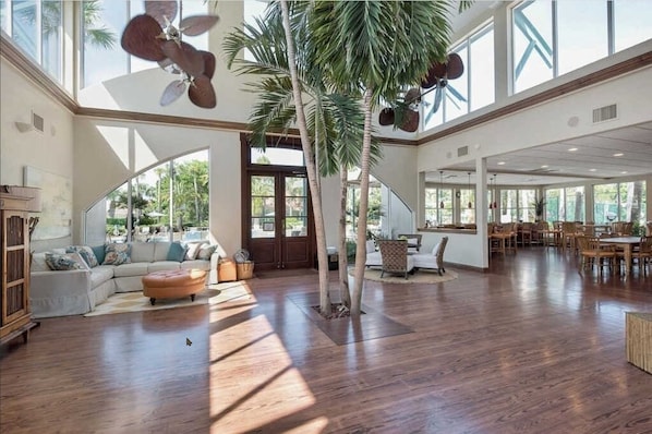 A spacious living area with a large wooden floor, a sofa set, a dining area with wooden tables and chairs, and a tall palm tree.