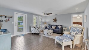 Ein geräumiges Wohnzimmer mit einem grauen Sofa, zwei gemusterten Hocker, einem Korbstuhl und einem Deckenventilator.