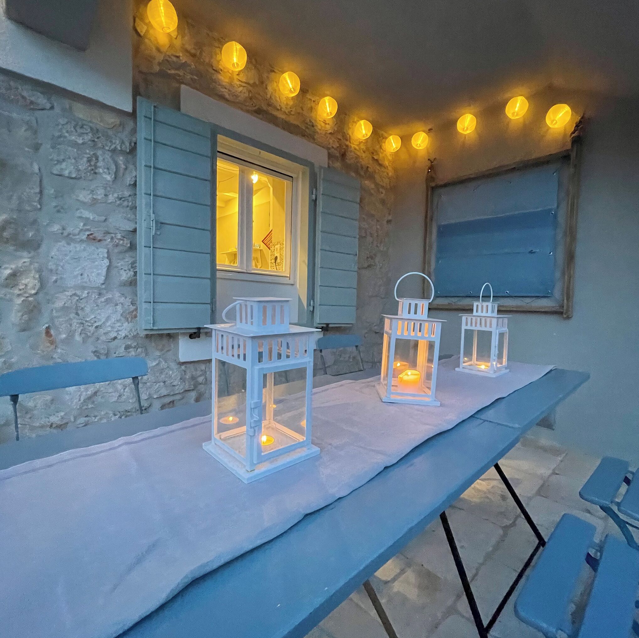A rustic outdoor seating area with a stone wall, a window with shutters, and two white lanterns on a table.