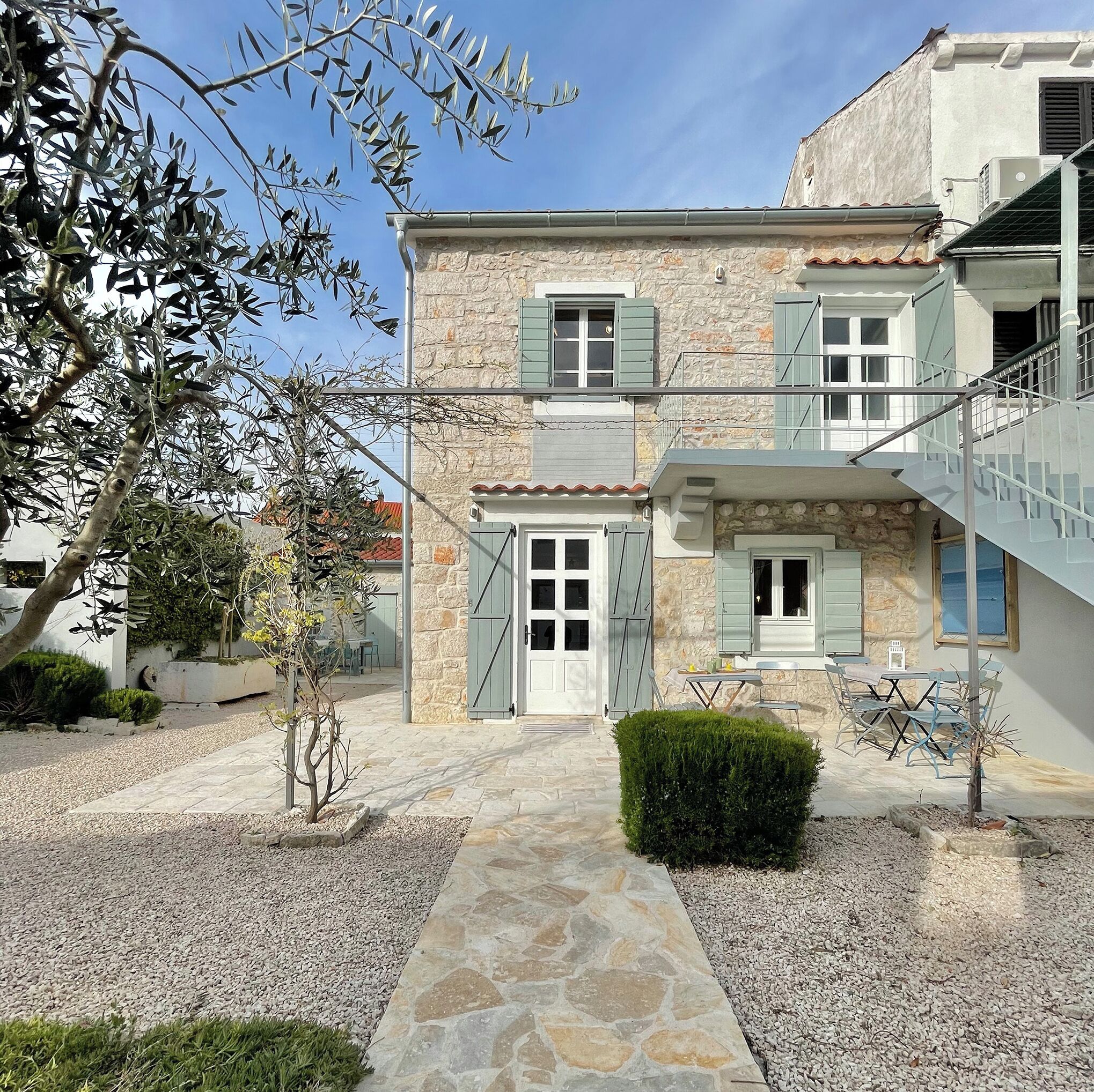 A two-story stone building with a central entrance, a small garden area with gravel and plants, and a staircase leading to an upper level.