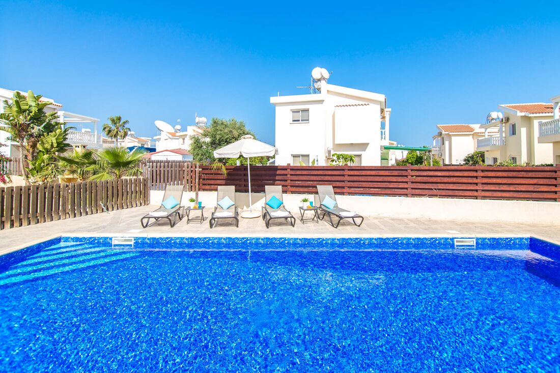 Une piscine avec des chaises longues, une clôture en bois et un bâtiment blanc en arrière-plan.