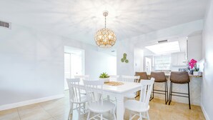Un luminoso comedor blanco con una araña redonda, sillas blancas y una cocina al fondo.