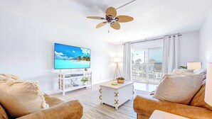 Una sala de estar moderna con un televisor de pantalla plana, un ventilador de techo, una mesa de centro blanca y un ventanal con cortinas.