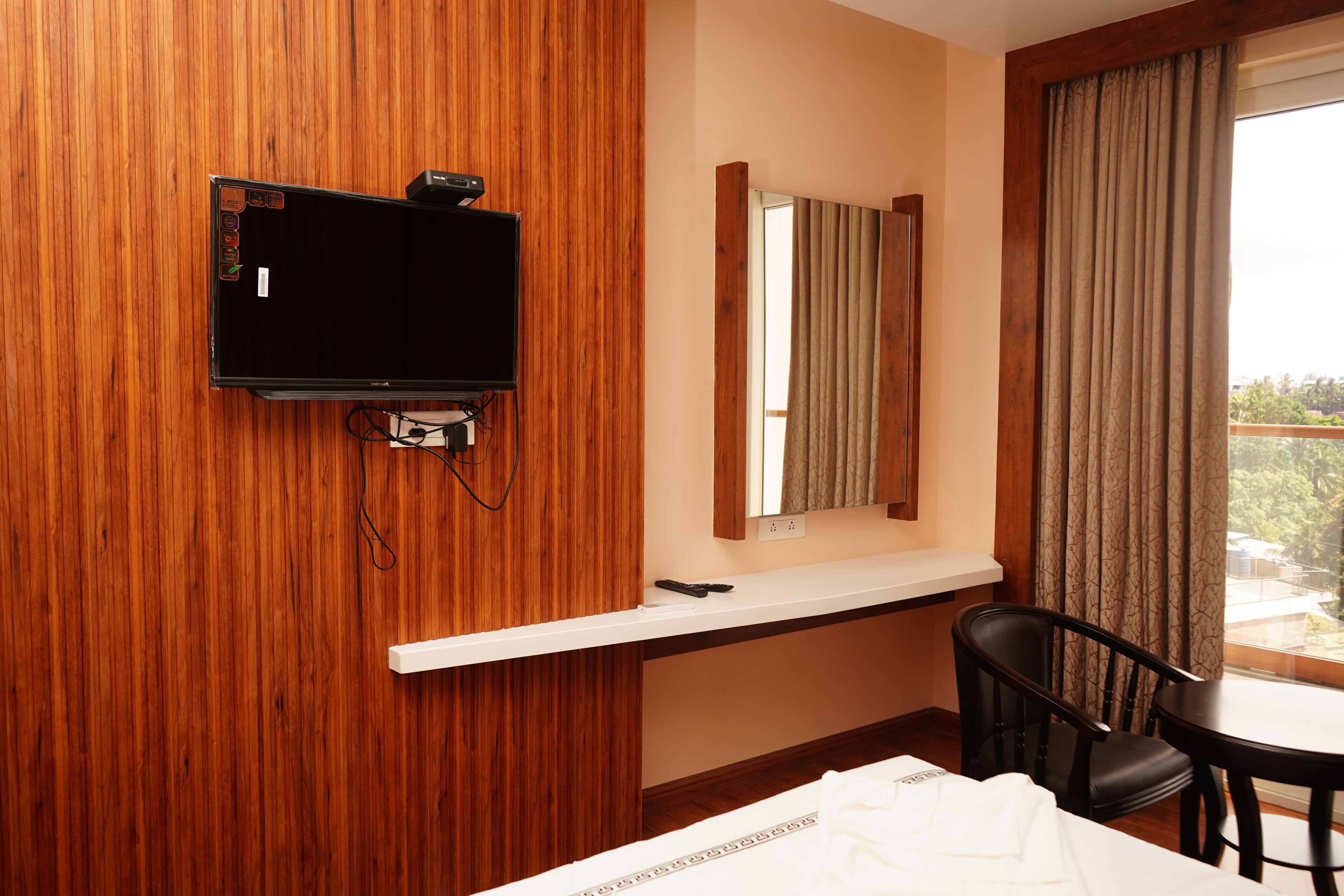Une chambre d’hôtel avec un mur en bois, un téléviseur à écran plat fixé au mur, une petite table avec deux chaises, un lit recouvert d’une couette blanche et une fenêtre avec des rideaux.