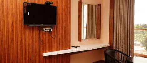 A hotel room with a wooden wall, a flat-screen TV mounted on it, a small table with two chairs, a bed with a white comforter, and a window with curtains.