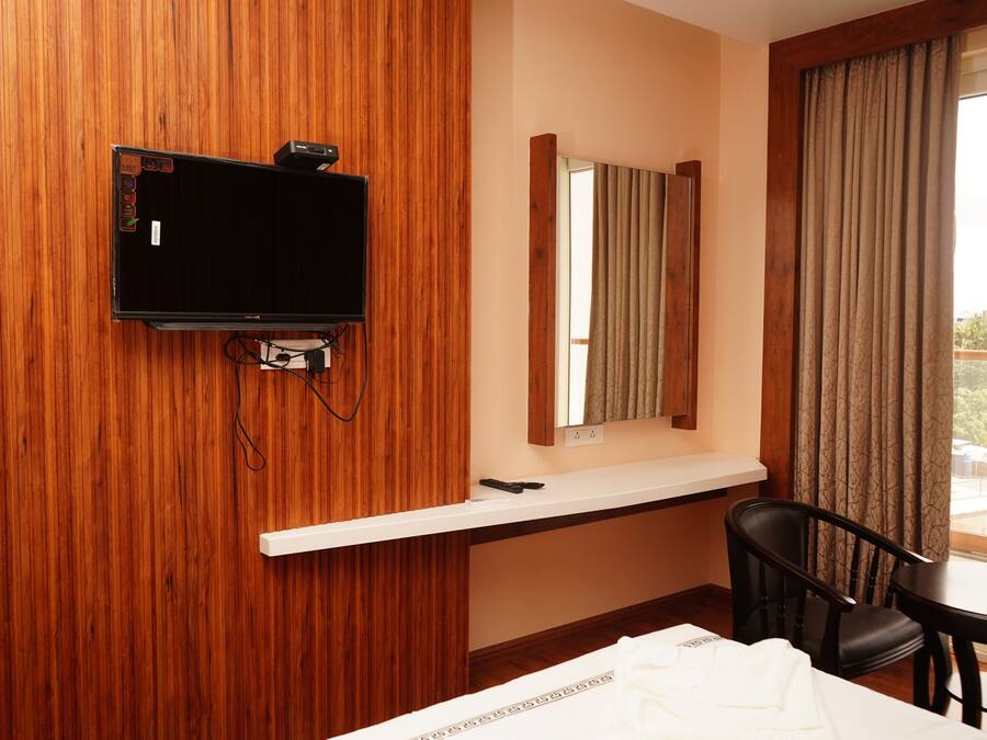 A hotel room with a wooden wall, a flat-screen TV mounted on it, a small table with two chairs, a bed with a white comforter, and a window with curtains.