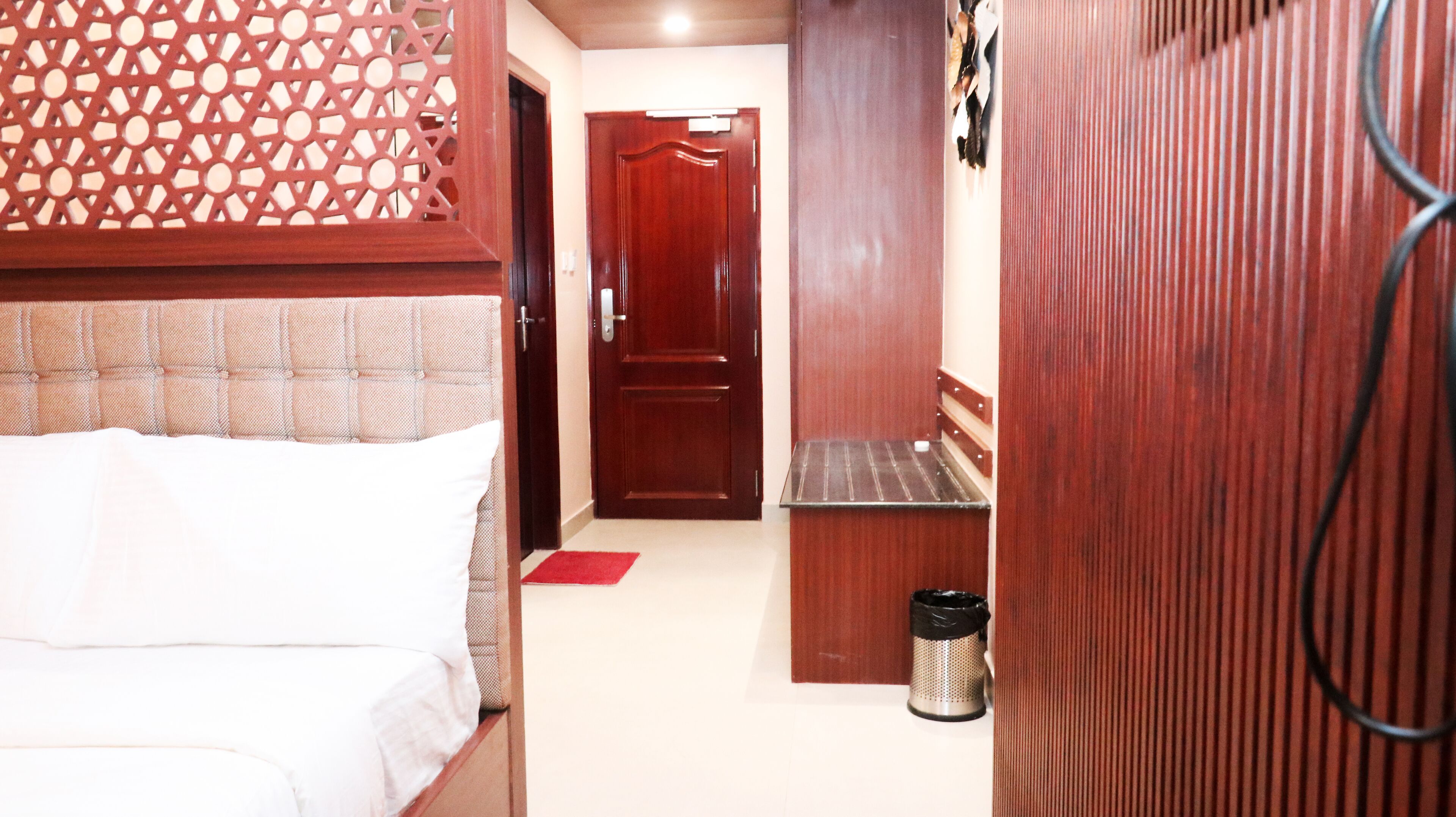A narrow hallway with a bed on the left, a door at the end, and a trash can on the right.