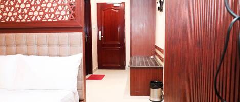 A narrow hallway with a bed on the left, a door at the end, and a trash can on the right.