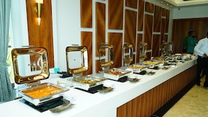 A buffet line with various food dishes, a wooden panel wall, and a person standing in the background. - Mansha Palace (Port Blair)