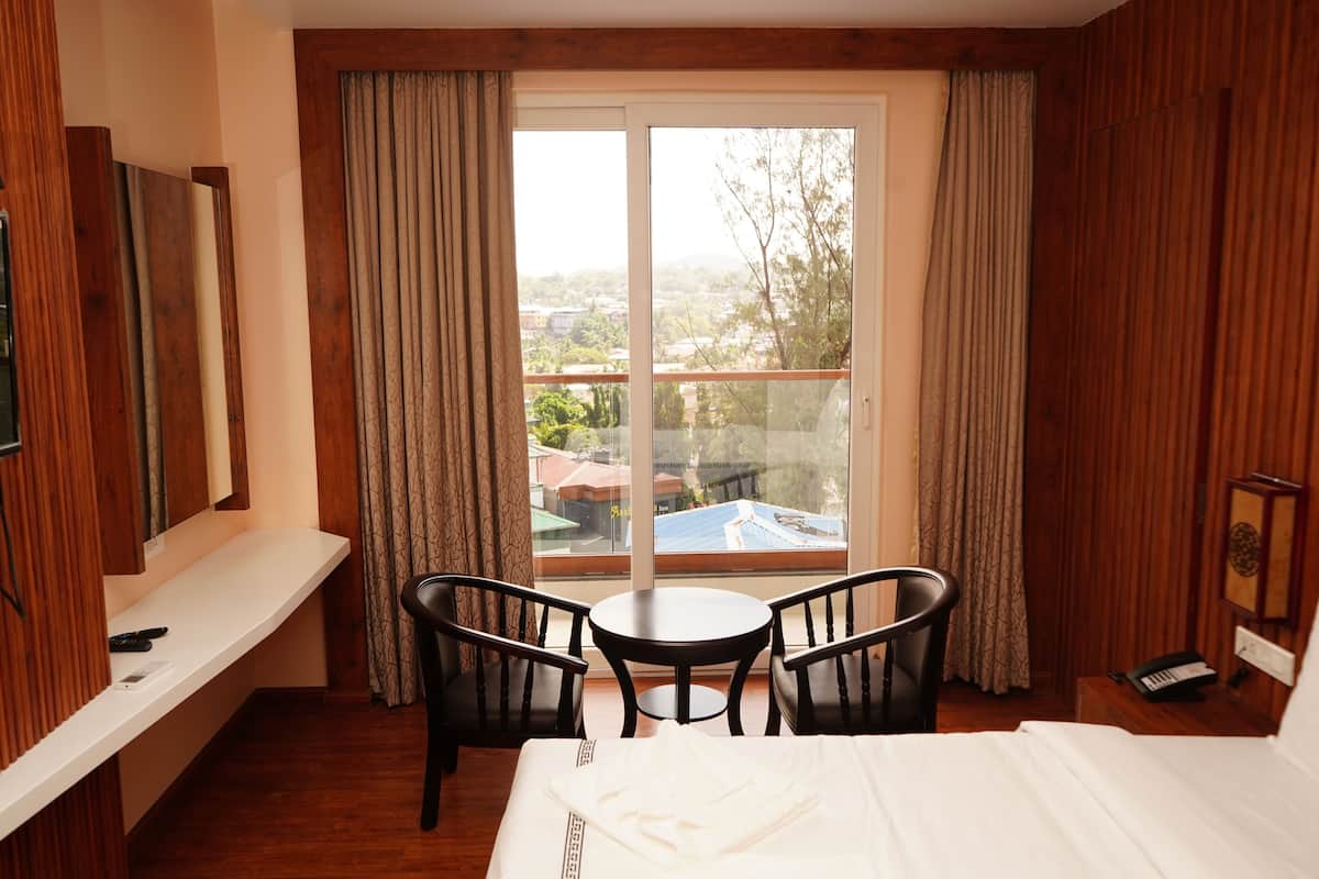 A hotel room with a bed, two chairs, a small table, a TV, and a balcony with curtains.