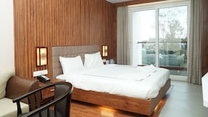 A modern hotel room with a wooden panel wall, a large bed with white bedding, a desk with a telephone, and a view of the outdoors through a large window. - Mansha Palace (Port Blair)