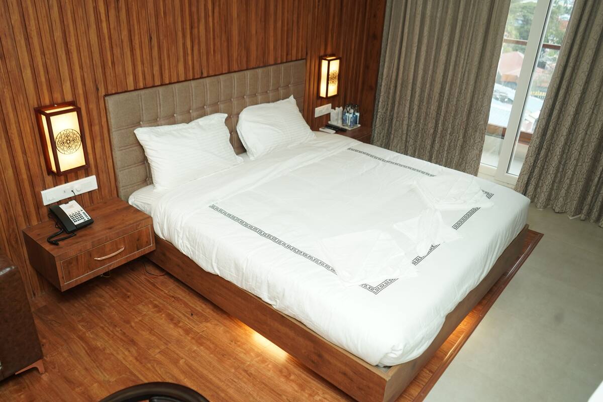 A hotel room with a large bed, wooden headboard, bedside lamps, a nightstand with a phone, and a window with curtains.
