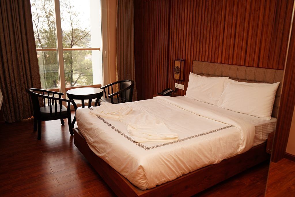 A hotel room with a large bed, a wooden headboard, a small table, two chairs, and a window with curtains.
