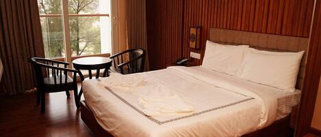 Habitación de hotel con una cama grande, cabecera de madera, una mesita, dos sillas y un ventanal con cortinas.