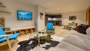 A modern living room with a glass coffee table, a blue armchair, a sofa, a mounted TV, and a kitchen in the background.