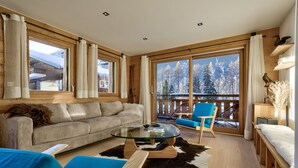 A cozy room with a sofa, a glass coffee table, a blue chair, and a view of snowy mountains through large windows.
