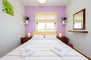 A bedroom with a bed, two bedside tables, a window with a blind, a mirror, and two wall-mounted shelves with plants.