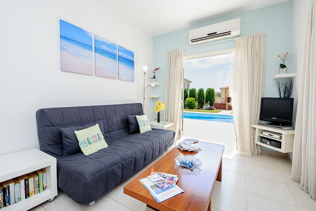 Ett modernt vardagsrum med en grå soffa, ett soffbord med tidningar, en tv på ett tv-bord och utsikt över en swimmingpool och grönområden utanför.