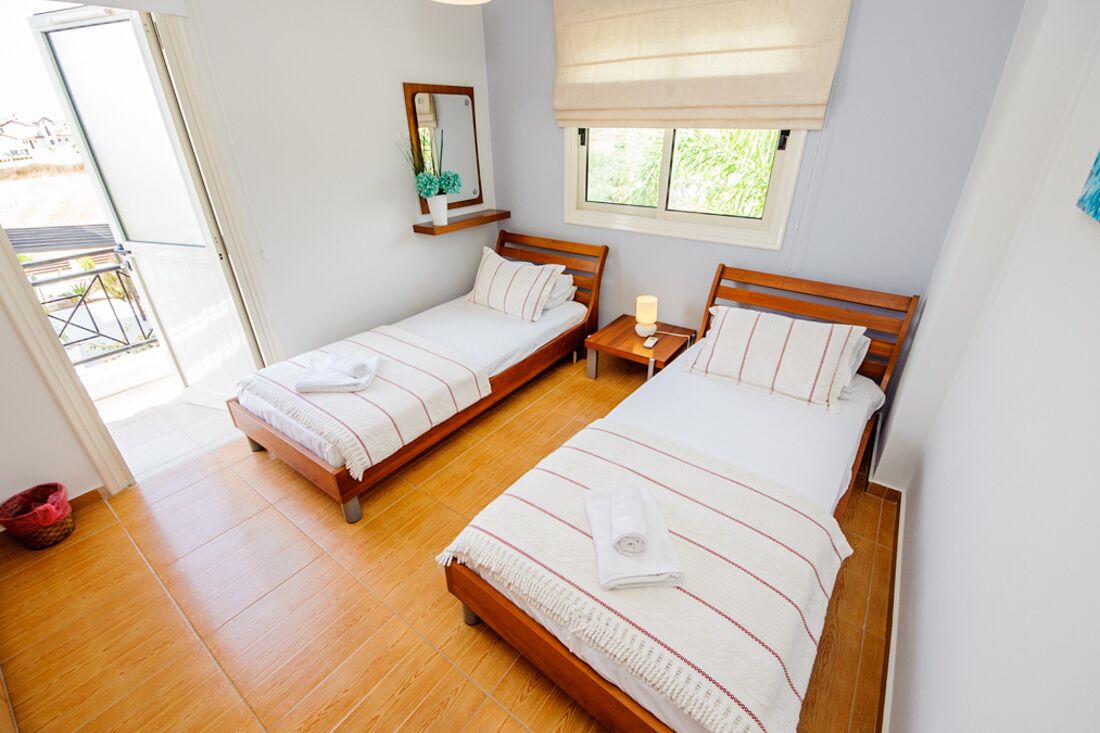 A bedroom with two wooden beds, a bedside table with a lamp, a window with a view of greenery, and a small shelf with a mirror and decorative items.
