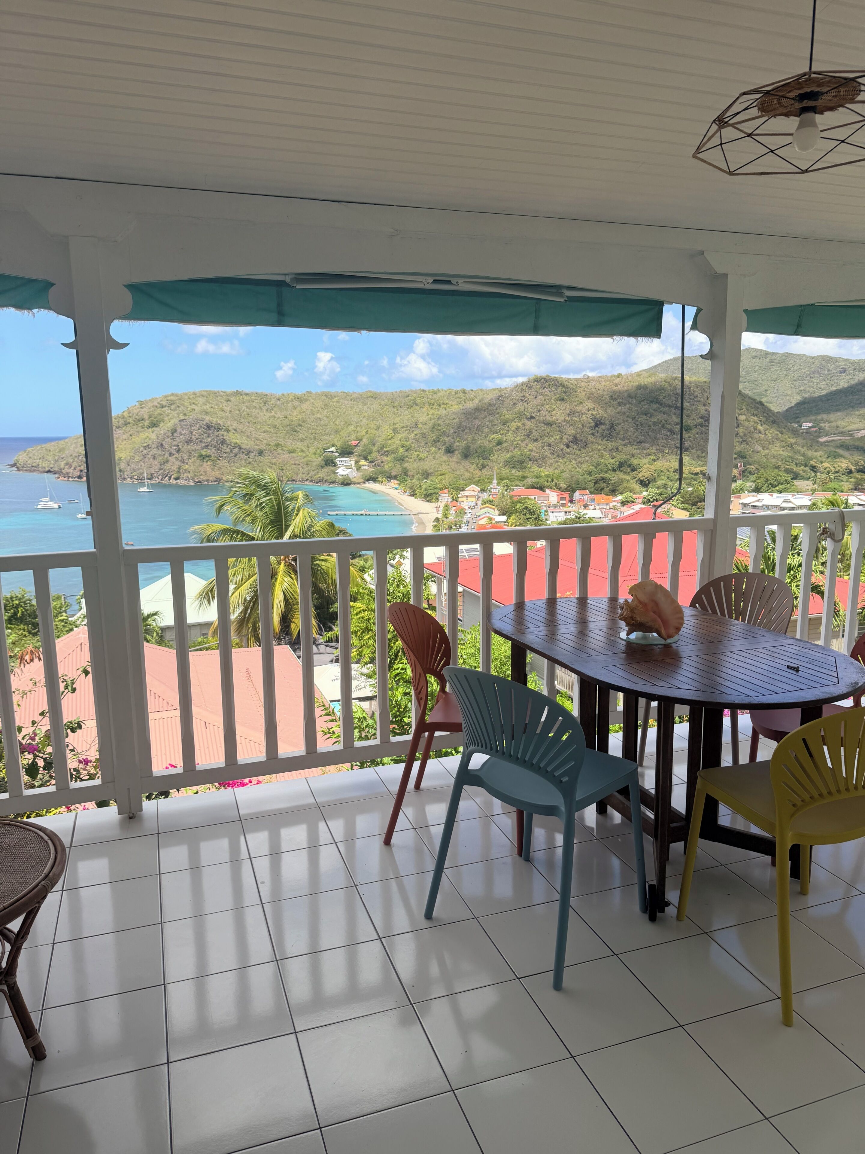 A covered outdoor area with a table and chairs, a view of a beach and mountains, and a tiled floor.