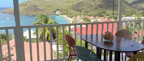 A covered outdoor area with a table and chairs, a view of a beach and mountains, and a tiled floor.