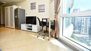 A modern living room with a flat-screen TV, a black office chair, a glass desk, and a large window with a city view. - Alia -Bright furnished studio near metro (Dubai)