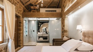 A bedroom with a large bed, a nightstand, a lamp, and a bathroom with a sink and mirror. - Chateau Anax Hotel (Rhodes)