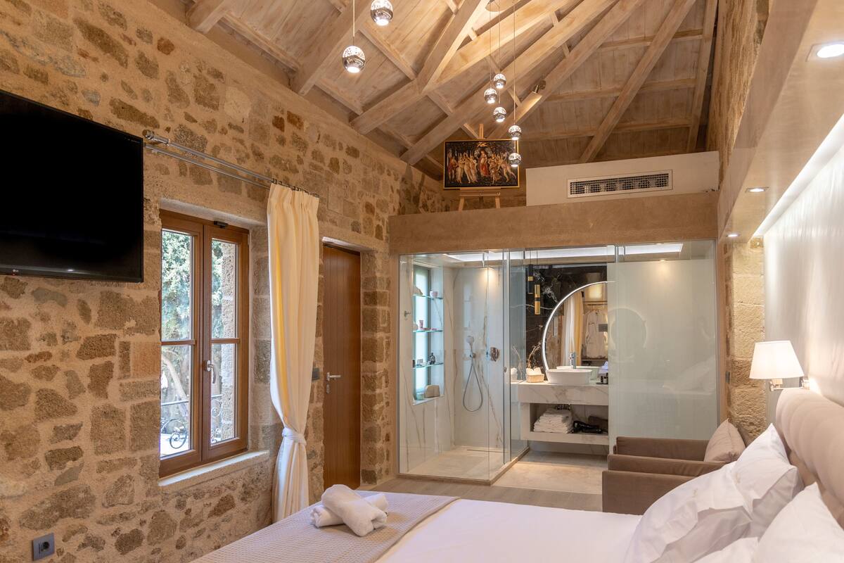 Ein Schlafzimmer mit Steinwänden, Holzboden und einem Bett mit weißer Bettwäsche. Ein Fernseher ist an der Wand montiert, und es gibt einen Duschbereich mit Glastüren.