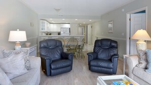 A living room with two black leather chairs, a white coffee table, and a grey sofa. A kitchen is visible in the background.