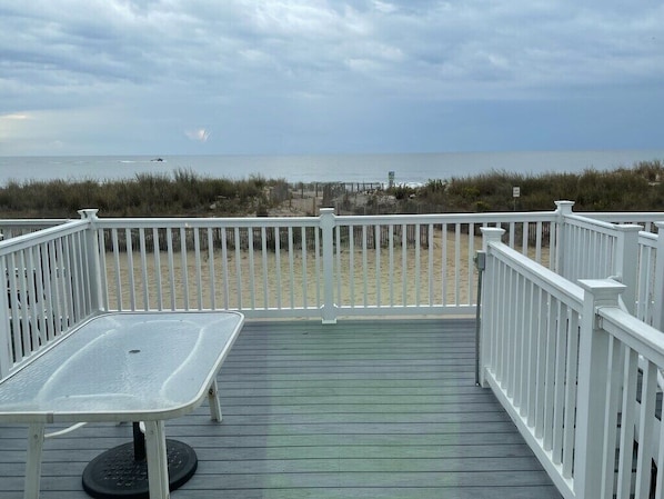 A wooden deck with a white railing, a metal table, and a view of the beach and ocean.