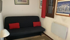 Un canapé noir avec des coussins rouges, un petit tabouret en bois, une lampe et un radiateur fixé au mur.