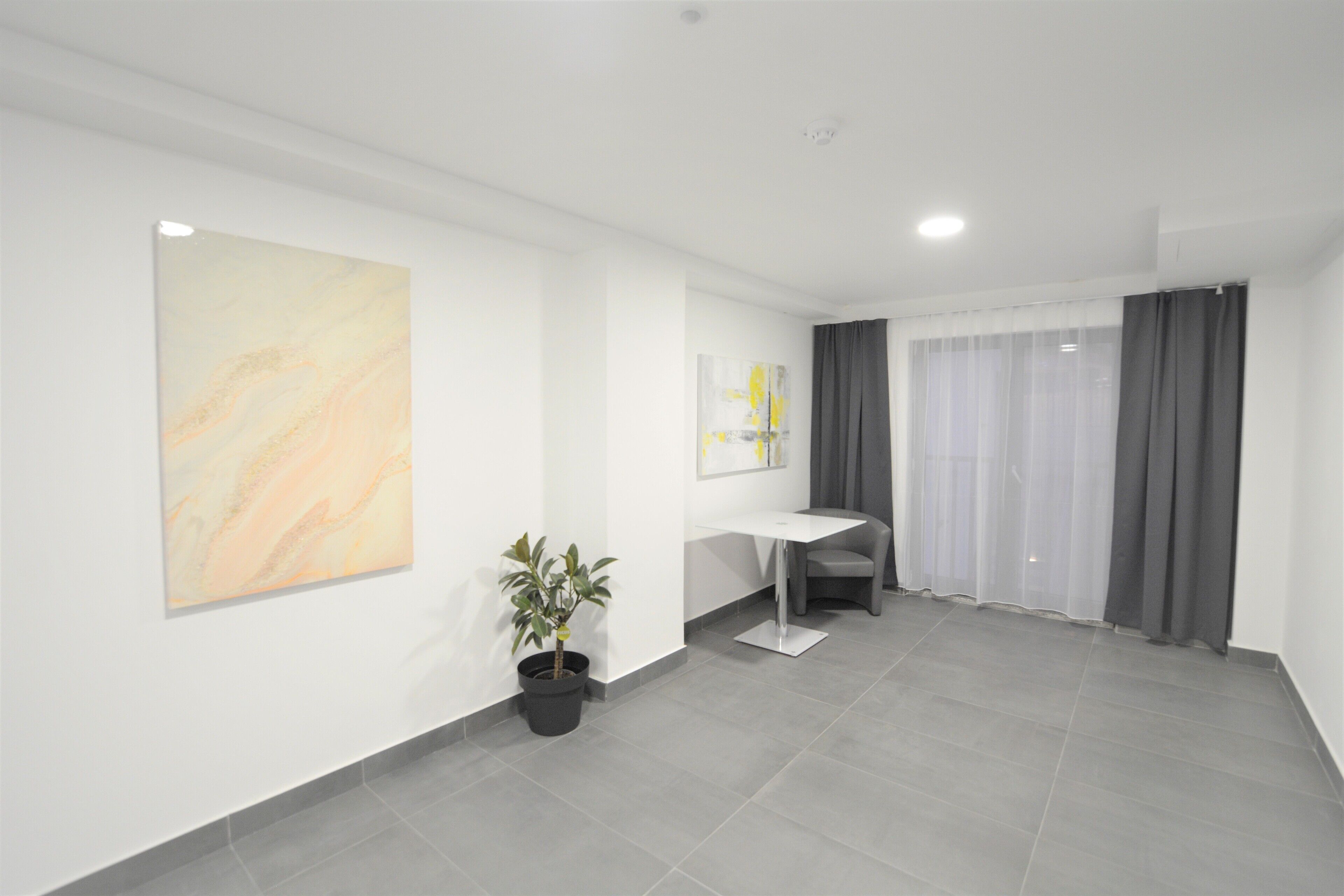 Une pièce moderne et minimaliste, avec une œuvre d’art en marbre au mur, une plante en pot, un bureau blanc et un rideau gris.
