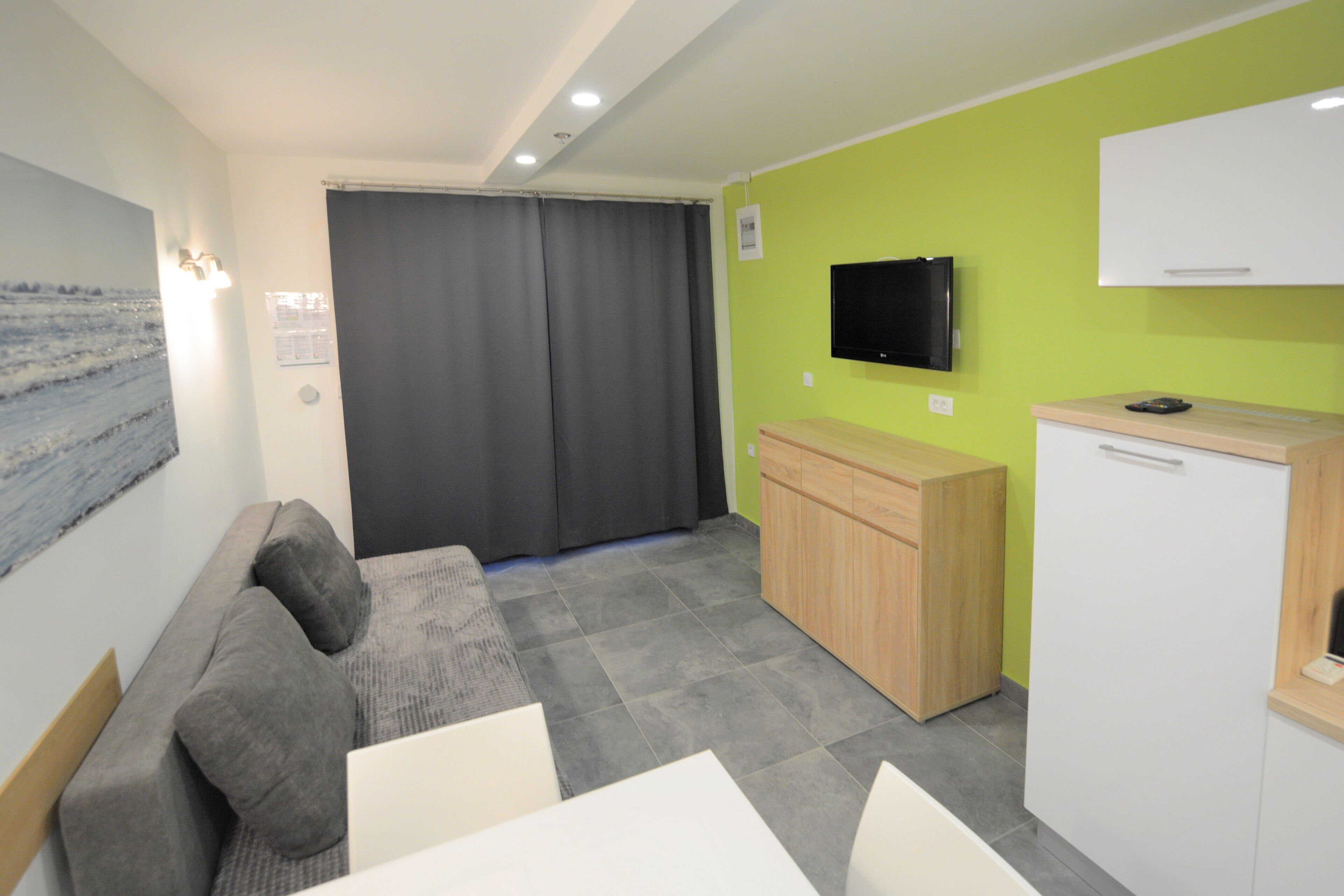 Una sala de estar moderna con un sofá gris, una mesita, un área de cocina con un refrigerador y un televisor montado en la pared.