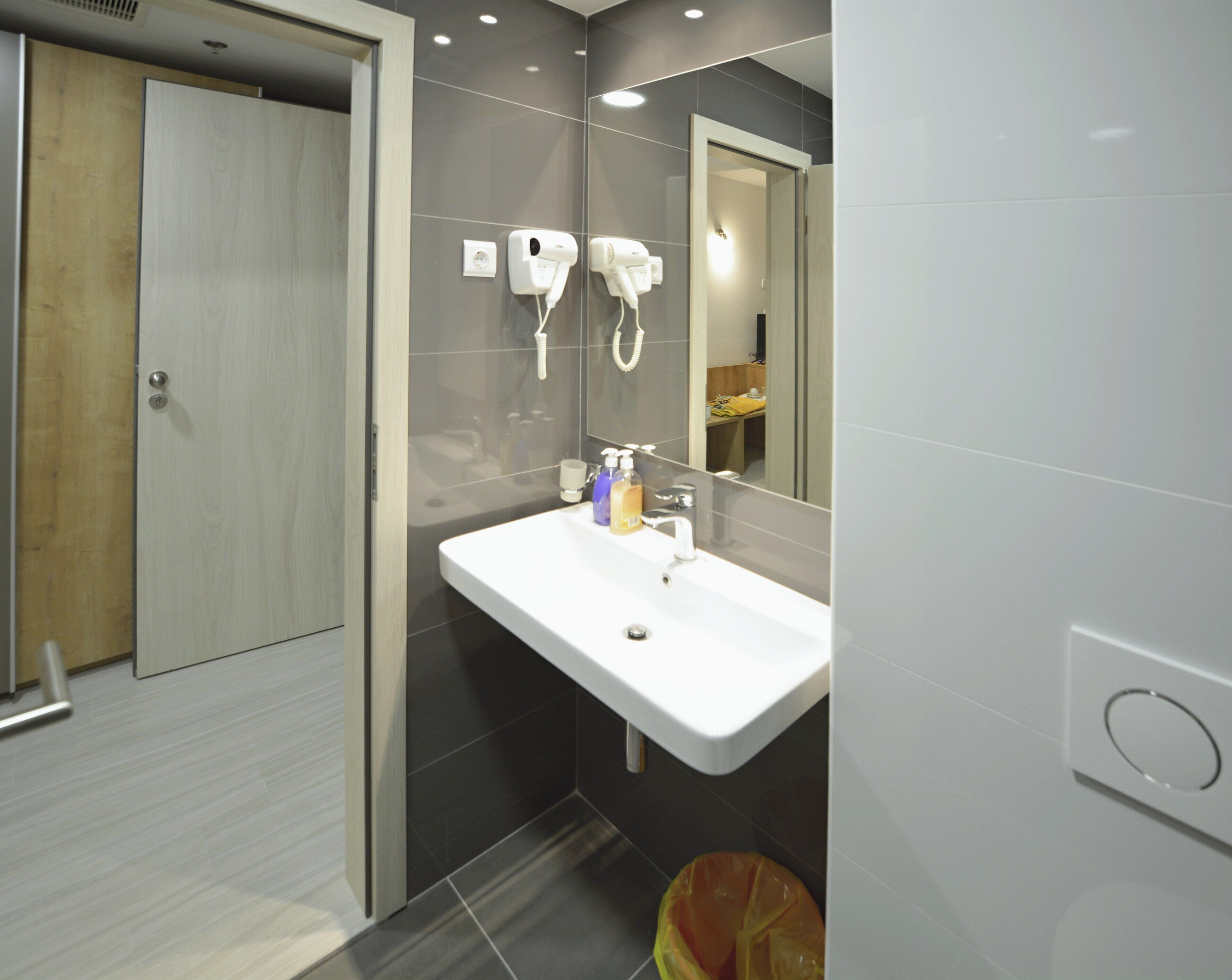 A modern bathroom with a white sink, two hair dryers, a mirror, and a trash bin.