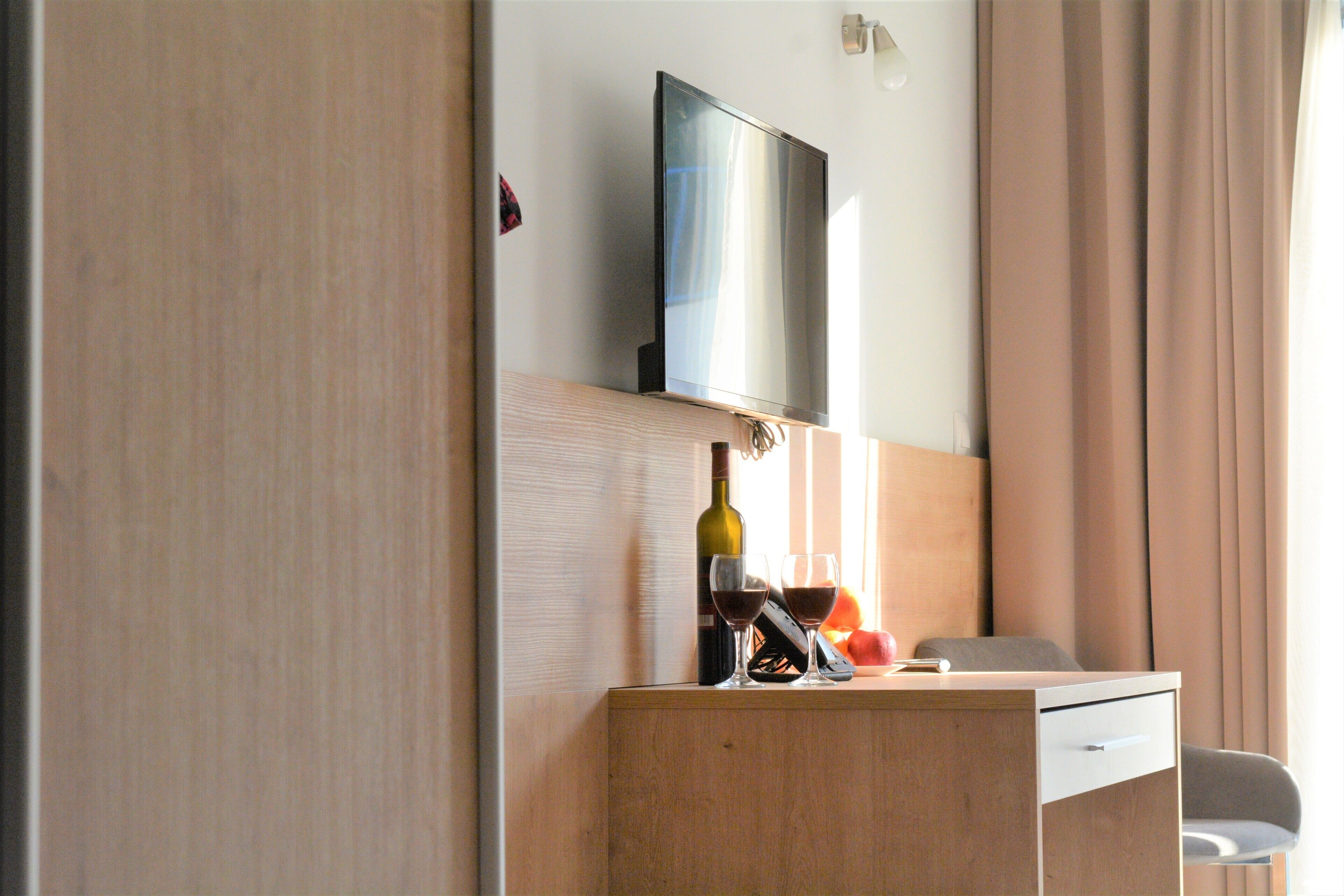 A hotel room with a wooden desk, a television mounted on the wall, a bottle of wine, and a glass on the desk.