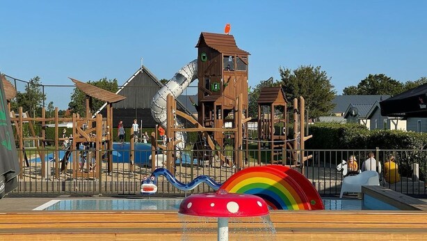 Zona infantil con tobogán, estructuras para escalar y una piscina con una fuente de agua con forma de hongo.