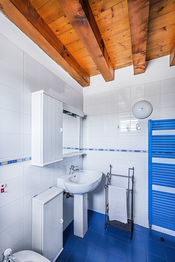 Baño con baldosas blancas, techo de madera, lavabo, espejo, toallero y cabina de ducha azul.