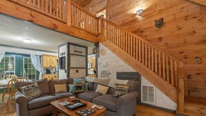 L’intérieur d’un chalet accueillant, avec un escalier en bois, un ensemble de canapés, un coin repas et une cuisine visible à travers une porte ouverte.