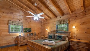 Une chambre à coucher située dans une cabane en bois, comprenant un lit, des tables de chevet, une commode, un ventilateur de plafond et une fenêtre avec des rideaux.