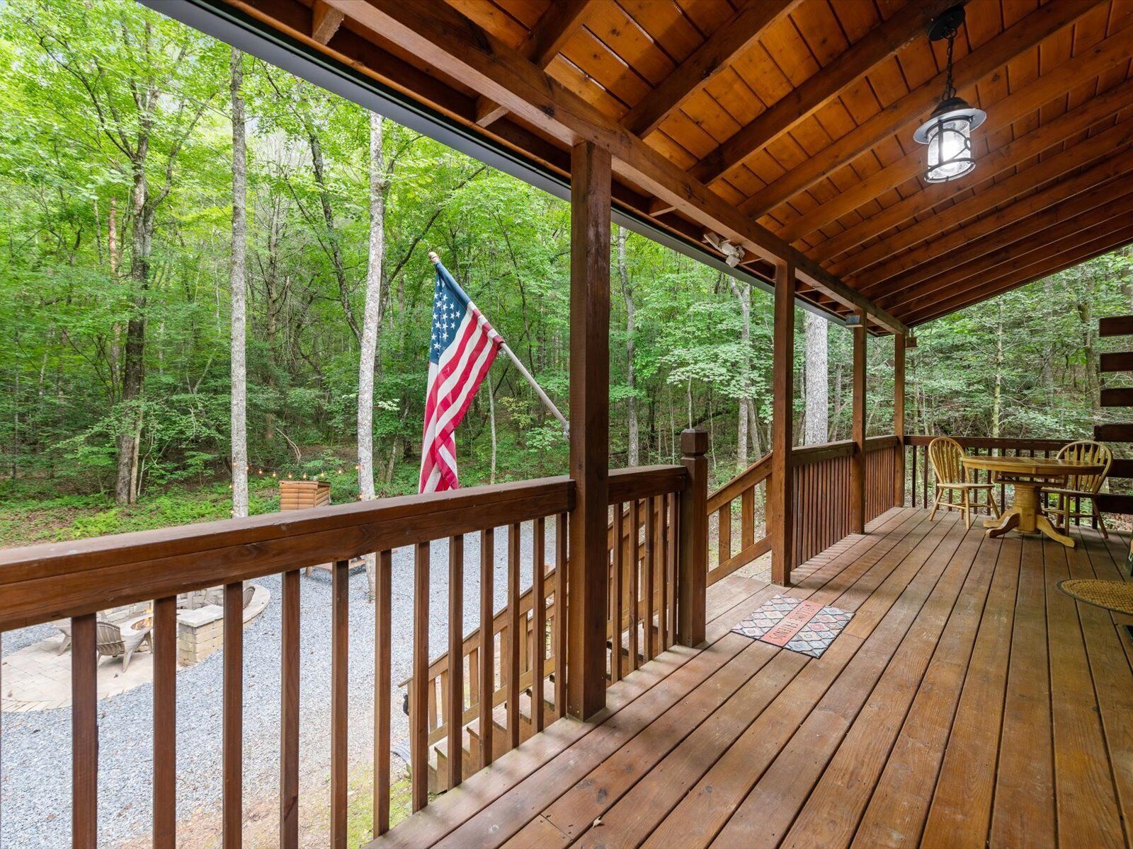 A wooden deck with a view of a forest, an American flag, and a table with chairs.