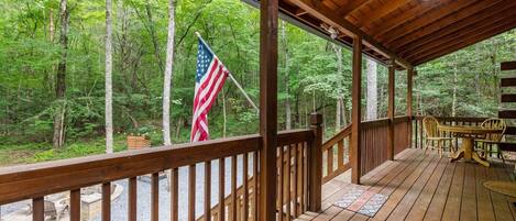 A wooden deck with a view of a forest, an American flag, and a table with chairs.
