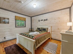 A cozy bedroom with a bed, a window, a wall with a 'be our guest' sign, and a small shelf. - Lazy Bear Cove - Blue Ridge, GA (Epworth)
