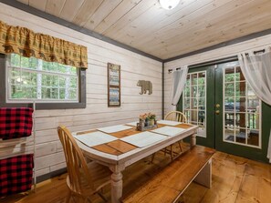 A cabin interior with a wooden dining table, chairs, a window with a view of trees, a door with glass panels, and a sign on the wall. - Lazy Bear Cove - Blue Ridge, GA (Epworth)