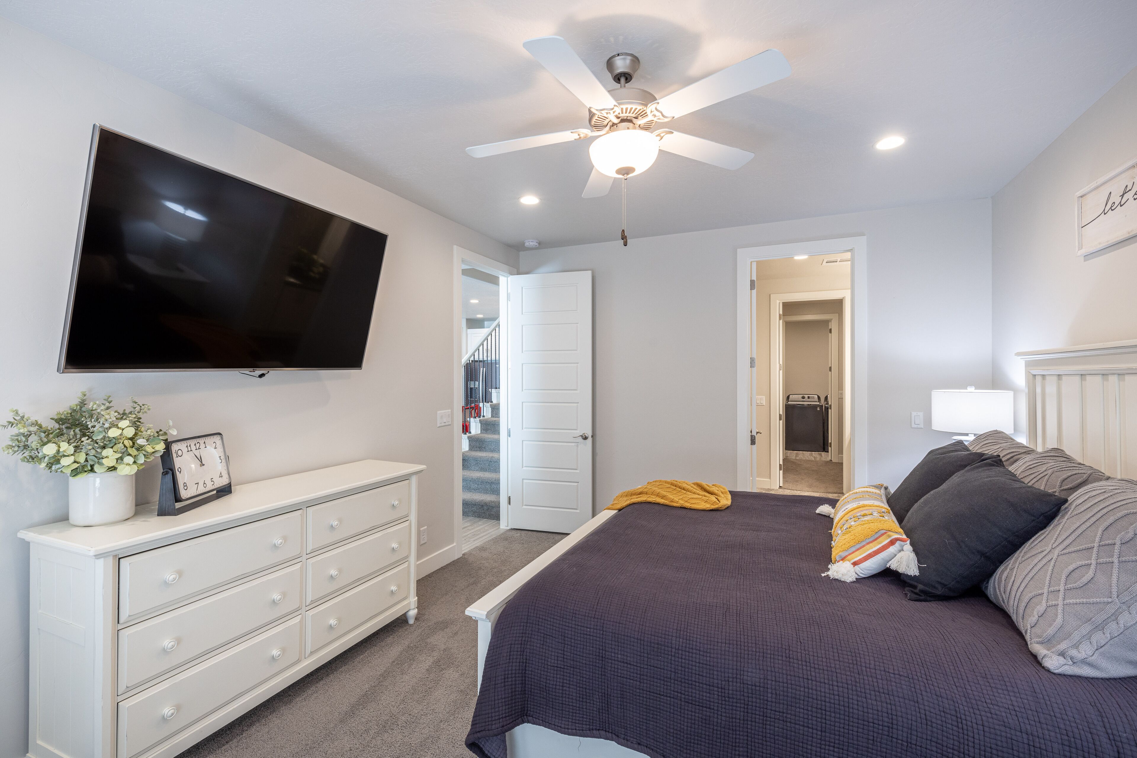 A bedroom with a bed, a dresser, a television, a ceiling fan, and a door leading to another room.